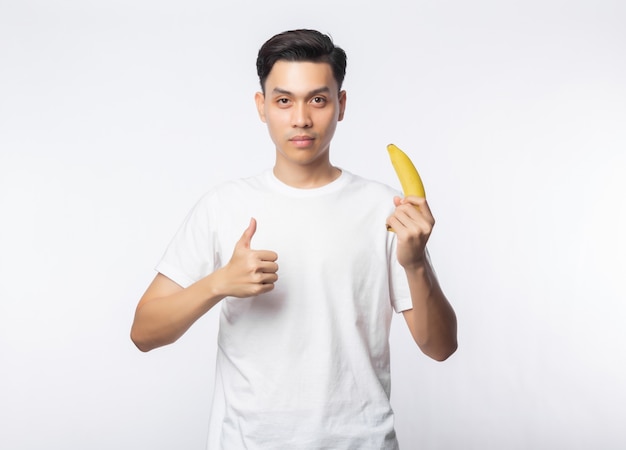 Jonge Aziatische mens in de witte banaan van de t-shirtholding en het tonen van duimen die omhoog op witte muur worden geïsoleerd