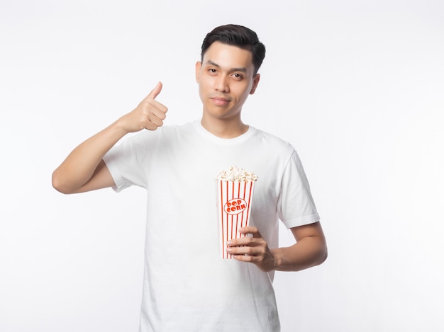 Jonge Aziatische mens in de witte popcorn van de t-shirtholding en het tonen van duimen die omhoog op witte muur worden geïsoleerd