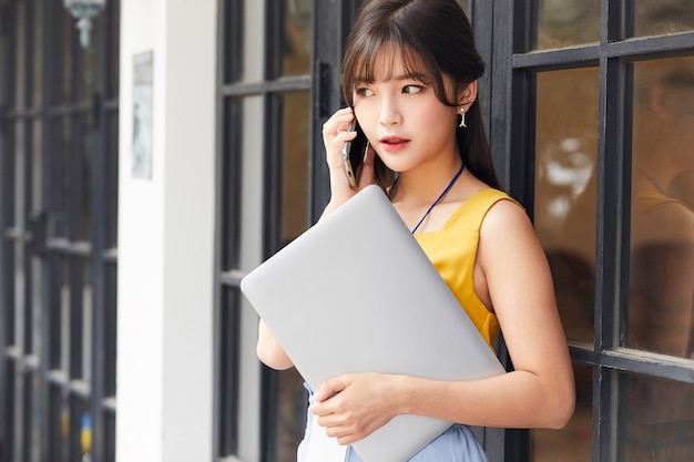 jonge Aziatische model zakenvrouw belt met haar smartphone terwijl ze een laptop op kantoor houdt