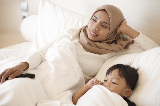 Jonge aziatische moeder en zoon spelen vrolijk op bed terwijl ze televisie kijken in een hotelkamer op vakantie