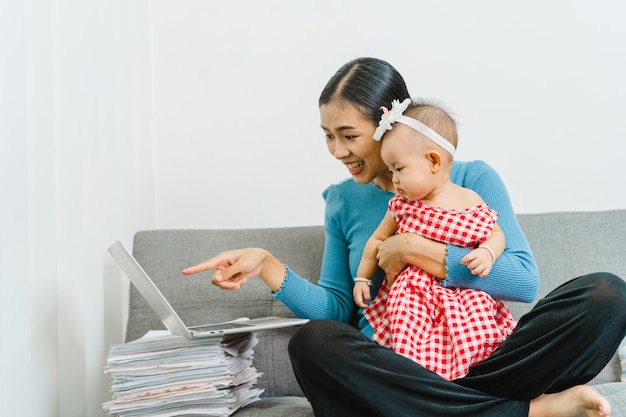 Jonge Aziatische moeder met schattige babydochter die samen op de bank in de woonkamer zit