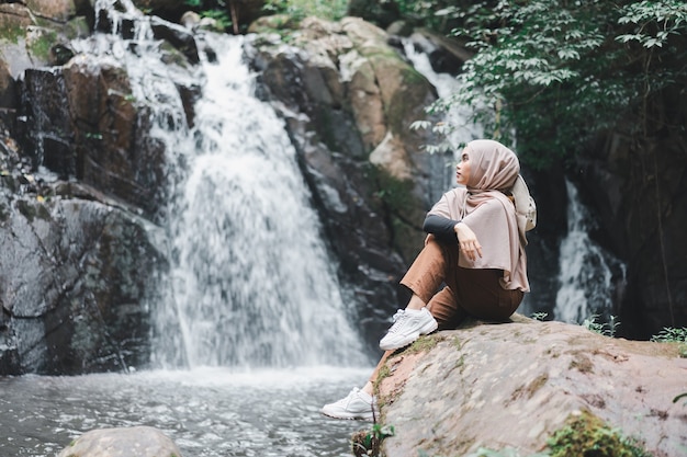 Jonge Aziatische moslim toeristenvrouw die bruine hijab draagt, zittend op de rots voor waterval.