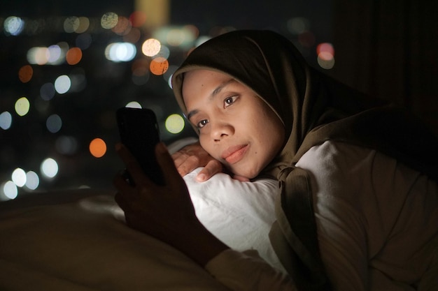 Jonge Aziatische moslimvrouw die een hoofddoek draagt terwijl ze in bed ligt en 's nachts een mobiele telefoon gebruikt