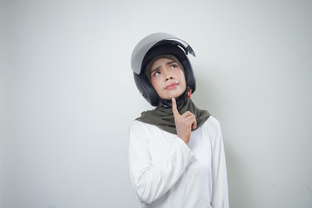 Jonge aziatische moslimvrouw die en denkt die motorhelm glimlacht die op witte achtergrond wordt geïsoleerd