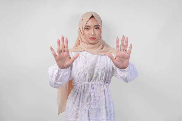 Foto jonge aziatische moslimvrouw in witte jurk en hijab die haar handpalm laat zien om een stopgebaar te maken met een serieuze uitdrukking