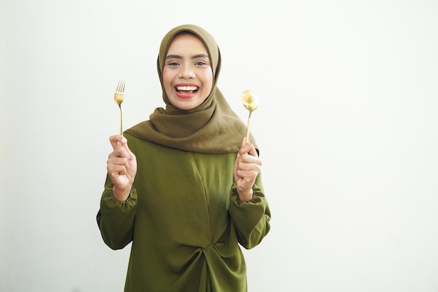 jonge Aziatische moslimvrouw met lepel en vork in haar mond en opzij kijkend dromerig heerlijk eten
