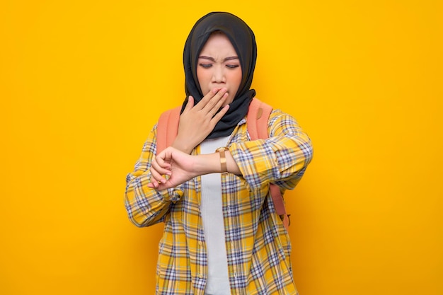 Jonge aziatische moslimvrouw student in casual kleding en rugzak tijd controleren op horloge gapende mond willen slapen geïsoleerd op gele achtergrond onderwijs school universiteit college concept