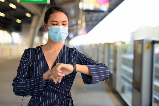 Jonge Aziatische onderneemster die met masker wacht en smartwatch controleert bij het skytrain-station