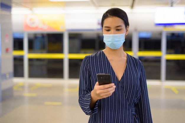 Jonge aziatische onderneemster met masker die telefoon met behulp van bij het metrostation