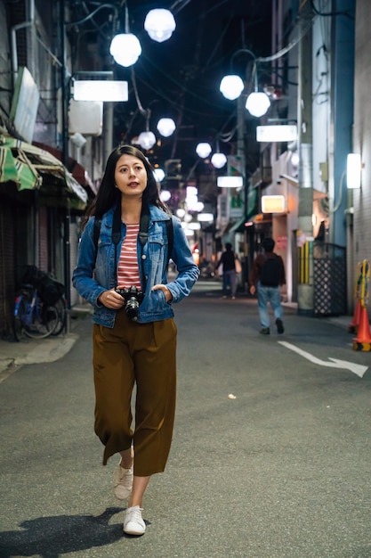 jonge aziatische reiziger die 's nachts teruggaat naar het hotel in osaka, japan. vrouwelijke toerist met camera avondje uit op de donkere straat. mooie vrouw met een glimlach die op de weg loopt met veel lichten aan.