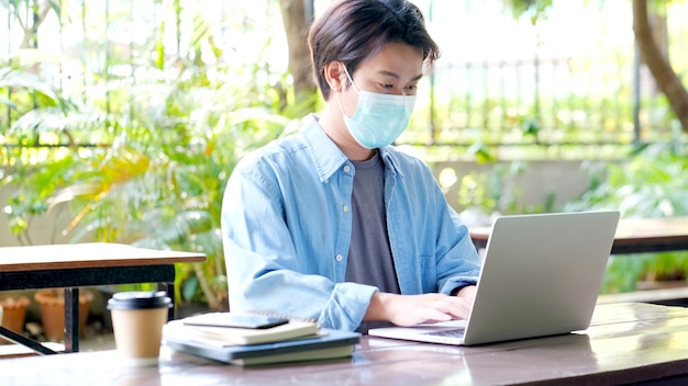 Jonge aziatische studentenmens die een beschermingsmasker draagt dat met laptop werkt