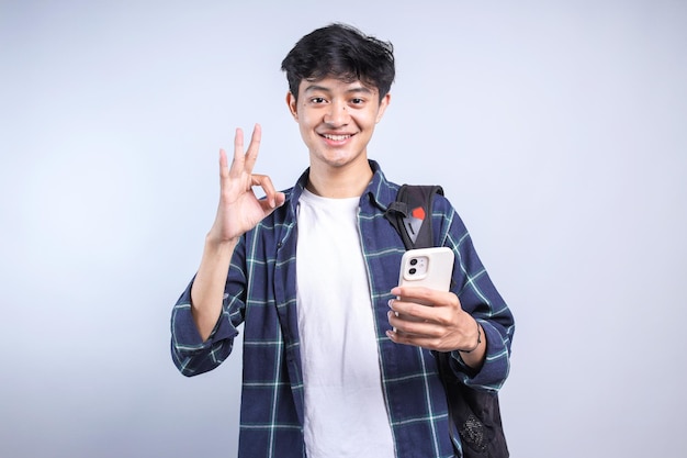 Jonge Aziatische universiteitsstudent draagt een rugzak met een smartphone terwijl hij een ok-teken toont met een gelukkige ex