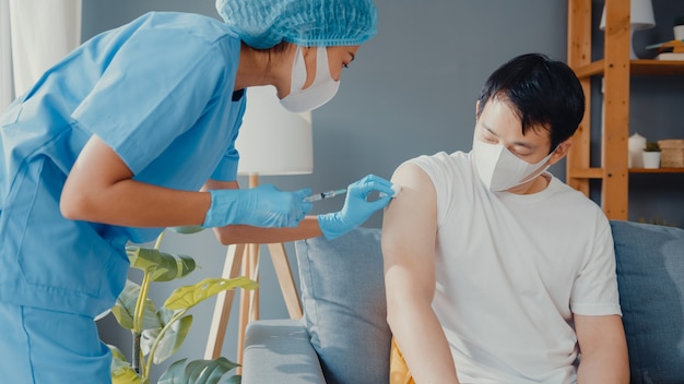 Jonge Aziatische verpleegster die Covid-19 of griep-antivirusvaccin geeft aan mannelijke patiënt, draagt gezichtsmasker ter bescherming tegen virussen