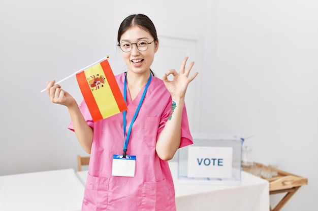 Jonge aziatische verpleegstersvrouw bij politieke campagneverkiezing die de vlag van spanje houdt die ok teken doet met vingers glimlachend vriendelijk gebarend uitstekend symbool