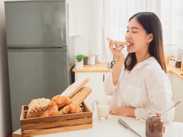 Jonge Aziatische vrouw die broodjes maakt en brood in keuken eet