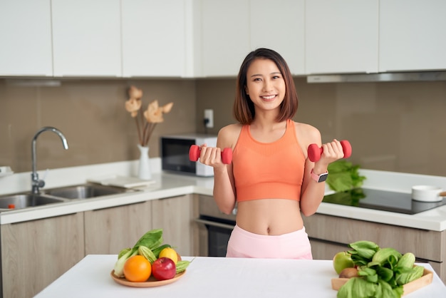 Jonge Aziatische vrouw die dumbell vasthoudt en sportieve kleding draagt in de keuken