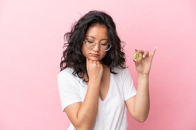 Jonge aziatische vrouw die een Bitcoin houdt die op roze achtergrond wordt geïsoleerd die twijfels heeft