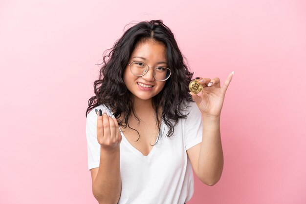 Jonge aziatische vrouw die een Bitcoin houdt die op roze achtergrond wordt geïsoleerd en geldgebaar maakt