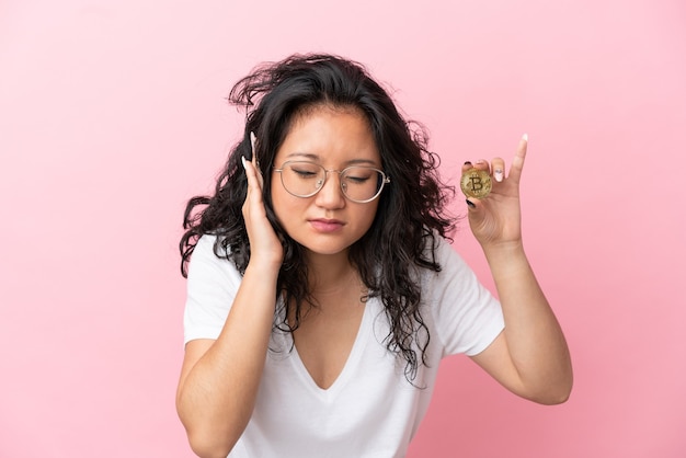 Jonge aziatische vrouw die een Bitcoin houdt die op roze gefrustreerde achtergrond wordt geïsoleerd en oren behandelt