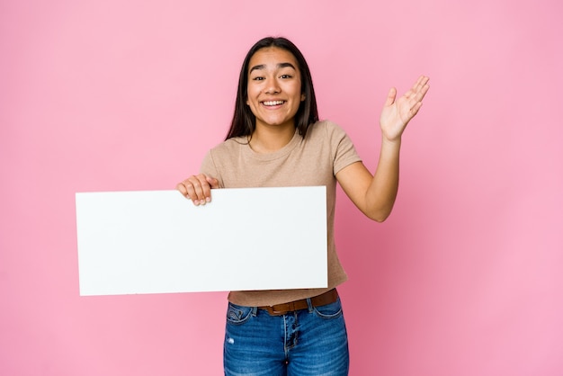 Jonge Aziatische vrouw die een blanco document voor wit houdt over geïsoleerde muur die een aangename verrassing ontvangt, opgewekt en handen opheft