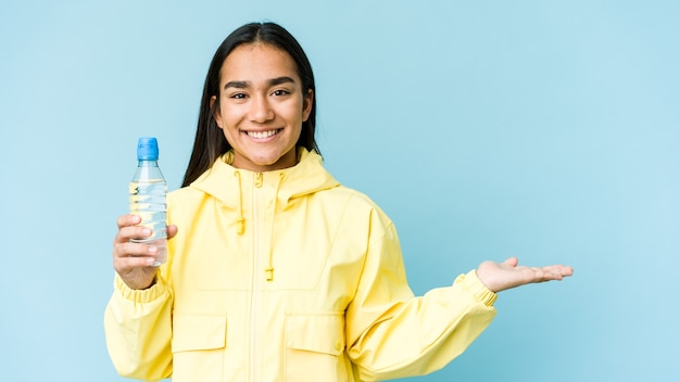 Jonge Aziatische vrouw die een fles water houdt dat op blauwe muur wordt geïsoleerd die een exemplaarruimte op een palm toont en een andere hand op taille houdt.