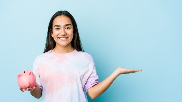 Jonge Aziatische vrouw die een roze bank houdt over geïsoleerde muur die een exemplaarruimte op een palm toont en een andere hand op taille houdt.