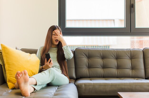 Jonge aziatische vrouw die geschokt, bang of doodsbang kijkt, gezicht bedekt met hand en tussen vingers gluurt