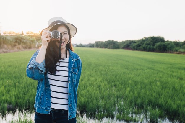 Jonge Aziatische vrouw die in hoed met camera glimlacht. Meisje die bij mooie aard met zonsondergang genieten van