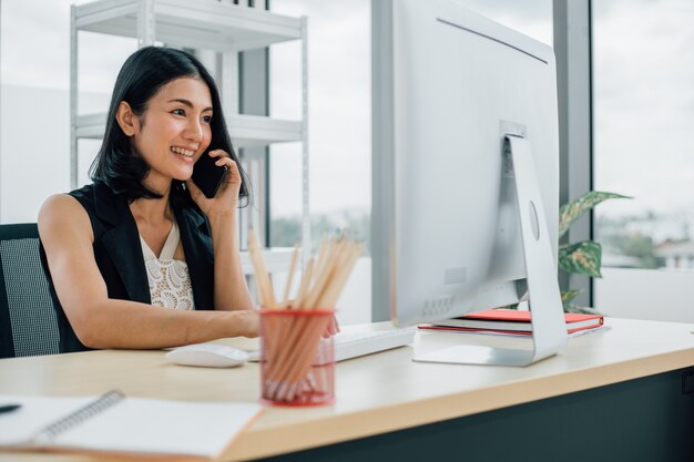 Jonge Aziatische vrouw die laptop computer met behulp van en telefonisch spreekt.