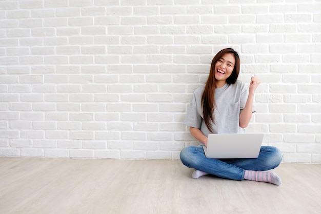 Jonge aziatische vrouw die laptop computerzitting voor witte bakstenen muur met behulp van