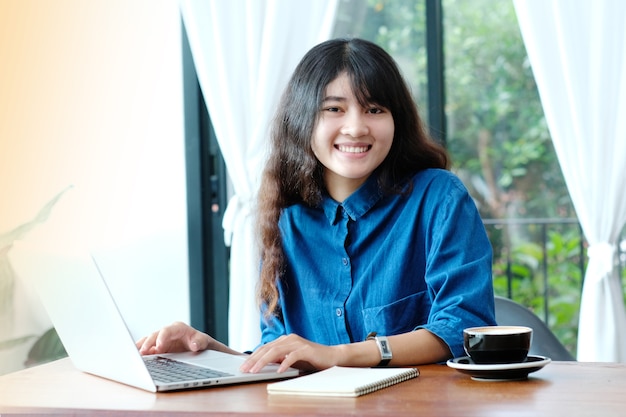 Jonge Aziatische vrouw die met laptop computer met het glimlachen gezicht in toevallige levensstijl werkt