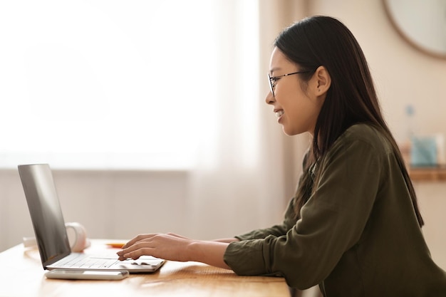 Jonge Aziatische vrouw die online werkt met behulp van laptop in de keuken