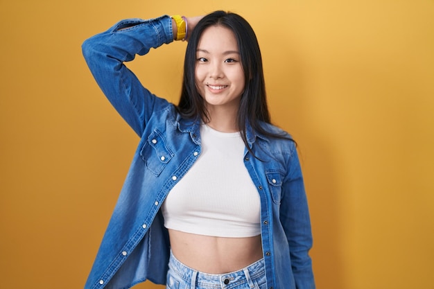 Jonge Aziatische vrouw die op een gele achtergrond staat, glimlacht zelfverzekerd, haar aanraakt met de hand omhoog, gestureert aantrekkelijk en modieus.