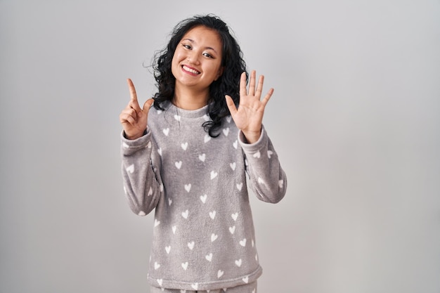 Jonge aziatische vrouw die pyjama draagt en met vingers nummer zeven naar boven wijst terwijl ze zelfverzekerd en gelukkig glimlacht