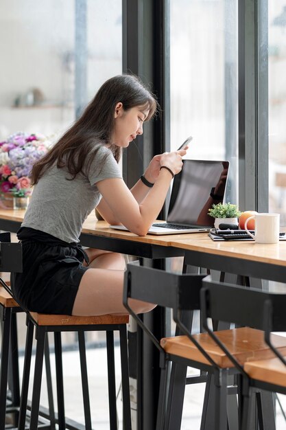 Jonge aziatische vrouw die smartphone en computer gebruikt terwijl ze in de co-werkruimte zit. verticale weergave.