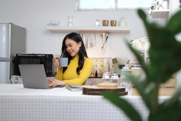 Jonge Aziatische vrouw drinkt koffie en werkt met een laptop in de keuken thuis