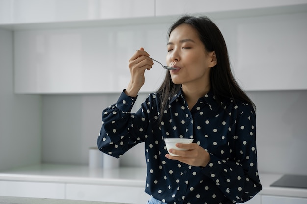 Jonge aziatische vrouw eet verse yoghurt als ontbijt in de keuken