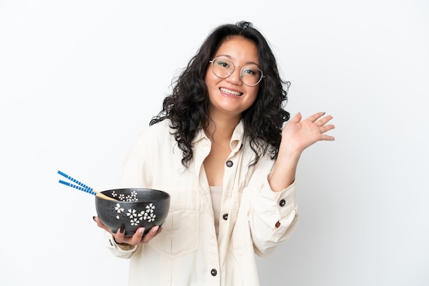 Jonge Aziatische vrouw geïsoleerd op een witte achtergrond die met de hand salueert met een gelukkige uitdrukking terwijl ze een kom noedels met eetstokjes vasthoudt