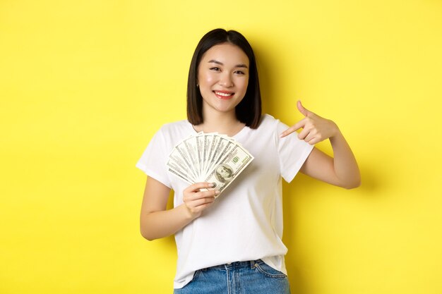Jonge Aziatische vrouw glimlachen, prijzengeld tonen, vinger wijzen naar dollars, staande over geel.