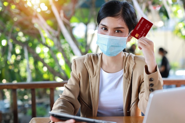 Jonge aziatische vrouw handen in casual kleding met beschermend masker voor gezondheidszorg, zittend in coffeeshop en tablet gebruiken en werken op laptopcomputer en creditcard tonen