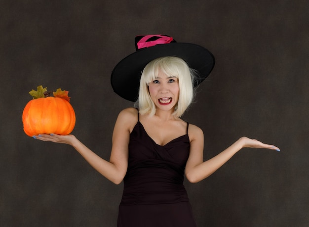 Jonge Aziatische vrouw in blonde pruik en heksenhoed met Halloween-pompoen die vrolijk lacht tijdens het feest op zwarte achtergrond
