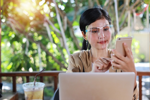 Jonge aziatische vrouw in casual kleding met gezichtsschild voor gezondheidszorg, zittend in de coffeeshop en gebruikend op smartphone en werkend op laptopcomputer. Nieuw normaal en social distancing concept