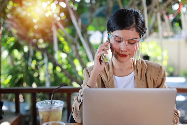 Jonge aziatische vrouw in casual kleding met gezichtsschild voor gezondheidszorg, zittend in de coffeeshop en gebruikend op smartphone en werkend op laptopcomputer. Nieuw normaal en social distancing concept