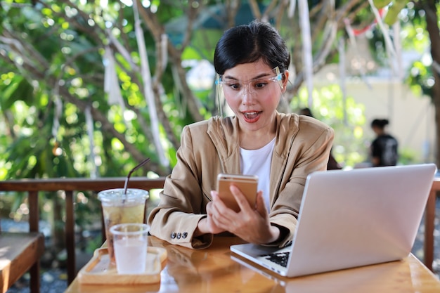 Jonge aziatische vrouw in casual kleding met gezichtsschild voor gezondheidszorg, zittend in de coffeeshop en gebruikend op smartphone en werkend op laptopcomputer. Nieuw normaal en social distancing concept