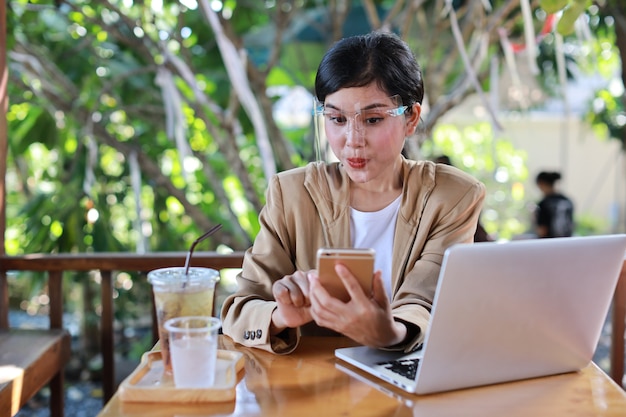 Jonge aziatische vrouw in casual kleding met gezichtsschild voor gezondheidszorg, zittend in de coffeeshop en gebruikend op smartphone en werkend op laptopcomputer. Nieuw normaal en social distancing concept
