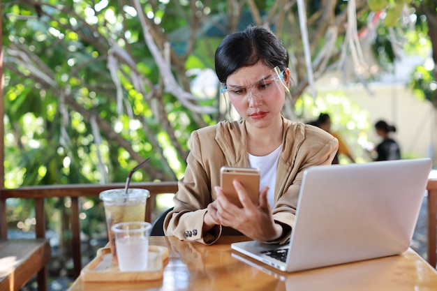 Jonge aziatische vrouw in casual kleding met gezichtsschild voor gezondheidszorg, zittend in de coffeeshop en gebruikend op smartphone en werkend op laptopcomputer. Nieuw normaal en social distancing concept