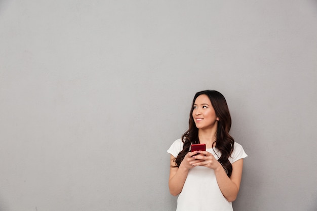 Jonge aziatische vrouw in casual t-shirt chatten of surfen op internet op mobiele telefoon en opzij kijken op copyspace, geïsoleerd over grijze muur