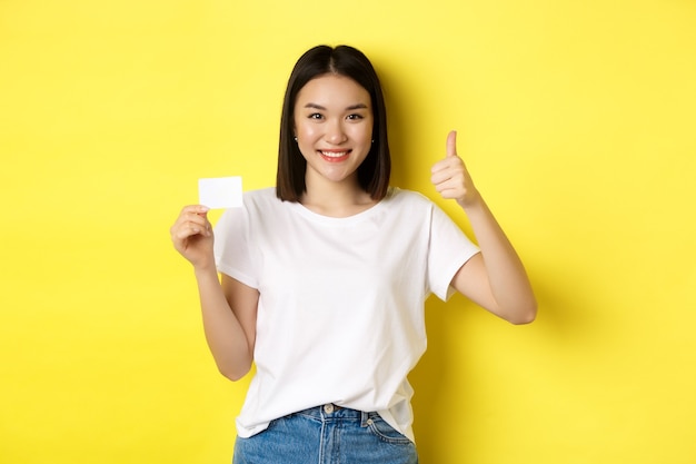 Jonge Aziatische vrouw in casual wit t-shirt met plastic creditcard en duim omhoog gebaar, goedkeuren en aanbevelen, glimlachend in de camera, gele achtergrond.