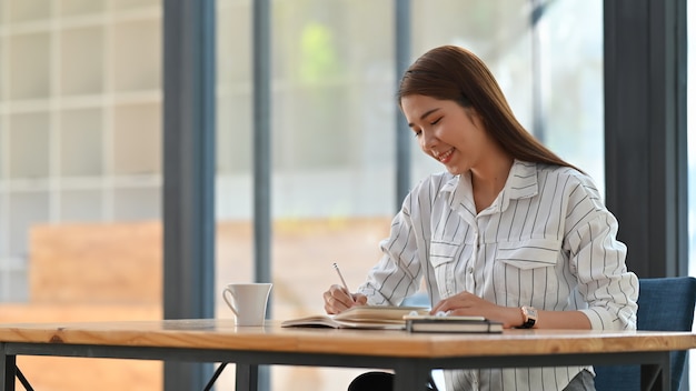 Jonge Aziatische vrouw in gestreept overhemd die / nota nemen nemen terwijl het zitten bij houten werkend bureau.