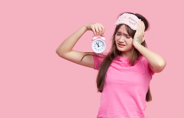 Jonge Aziatische vrouw in het masker die van het slaapoog geeuwen en wekker houden die op roze wordt geïsoleerd. Slapeloze nacht